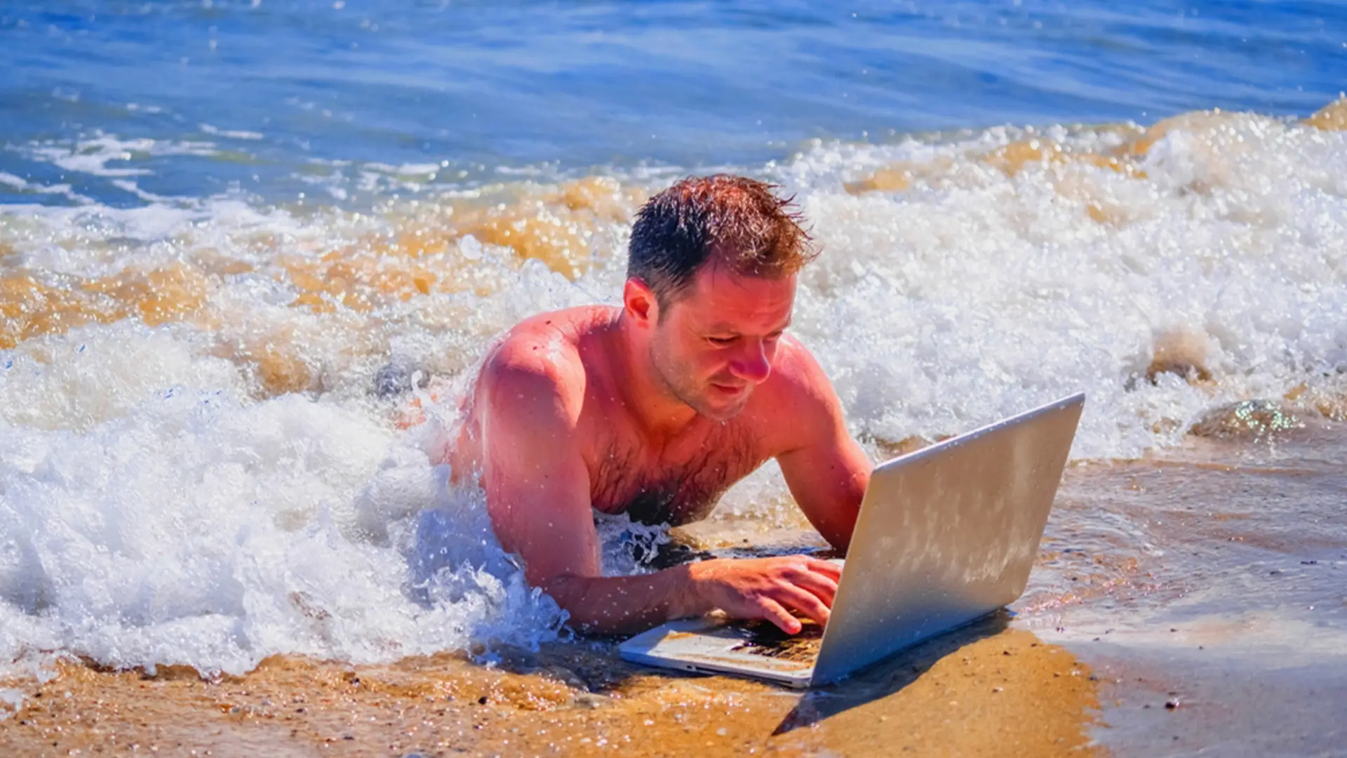 Homme utilisant un ordinateur portable en bord de mer, allongé dans les vagues.